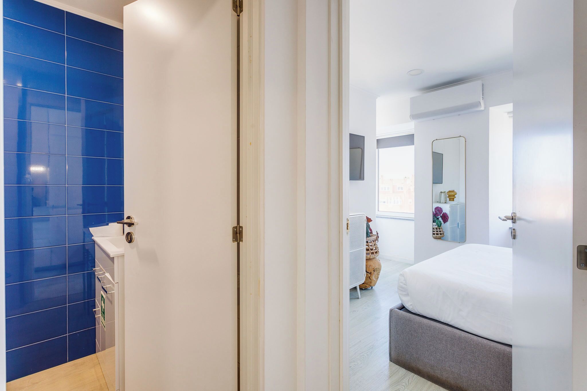 A hotel room with a white door, a blue tiled wall, a white sink, a bed with a grey headboard, and a mirror on the wall.