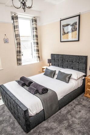 A neatly made bed with a dark headboard, a window with plaid curtains, and a framed picture on the wall.
