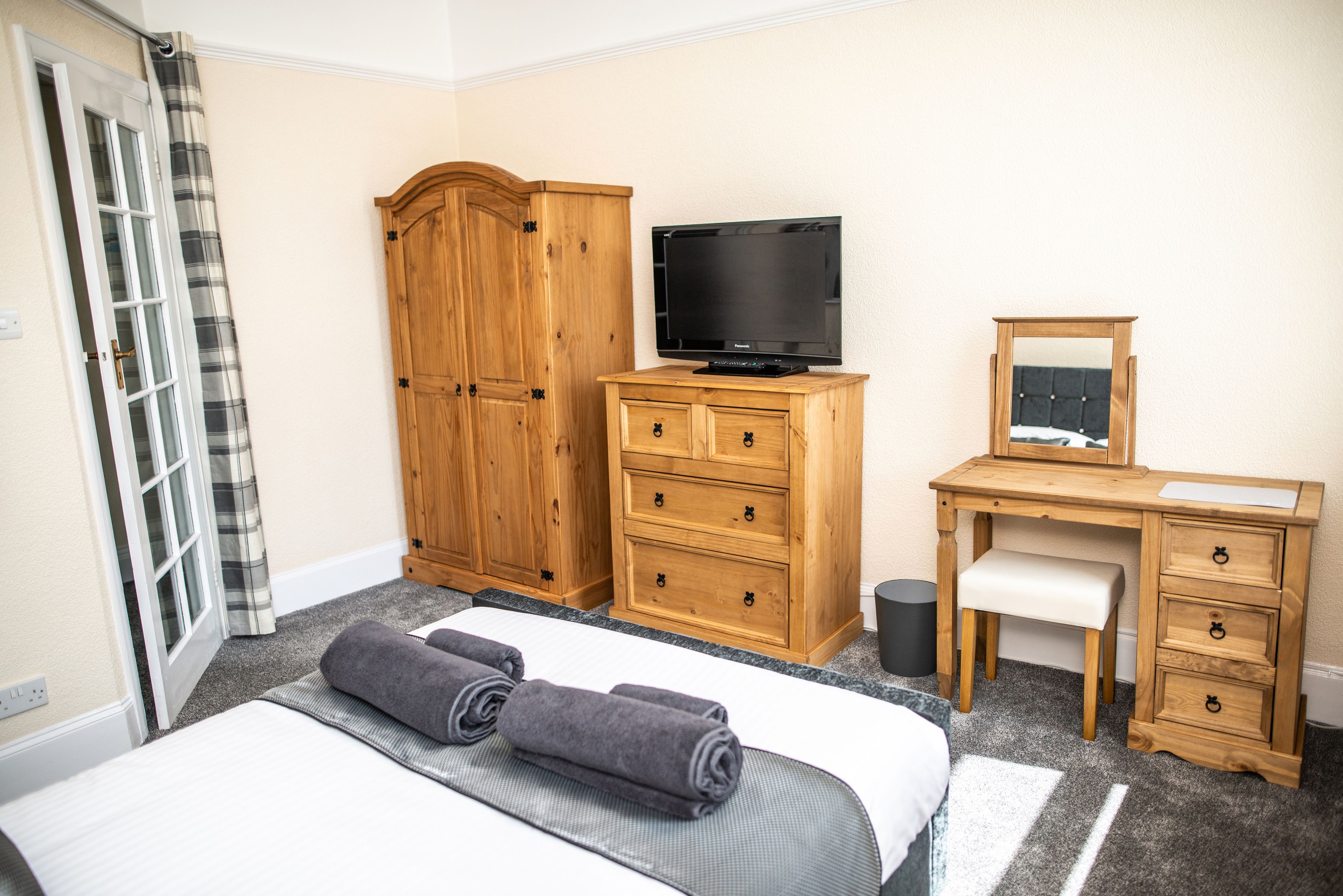 A hotel room with a bed, a wooden wardrobe, a desk with a chair, a television, and a mirror.