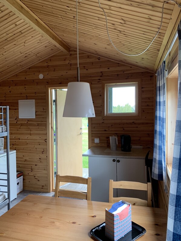 A wooden cabin interior with a dining table, chairs, a kitchen area, and a window. - Stuga på Nässjö Golfklubb (Nassjo)