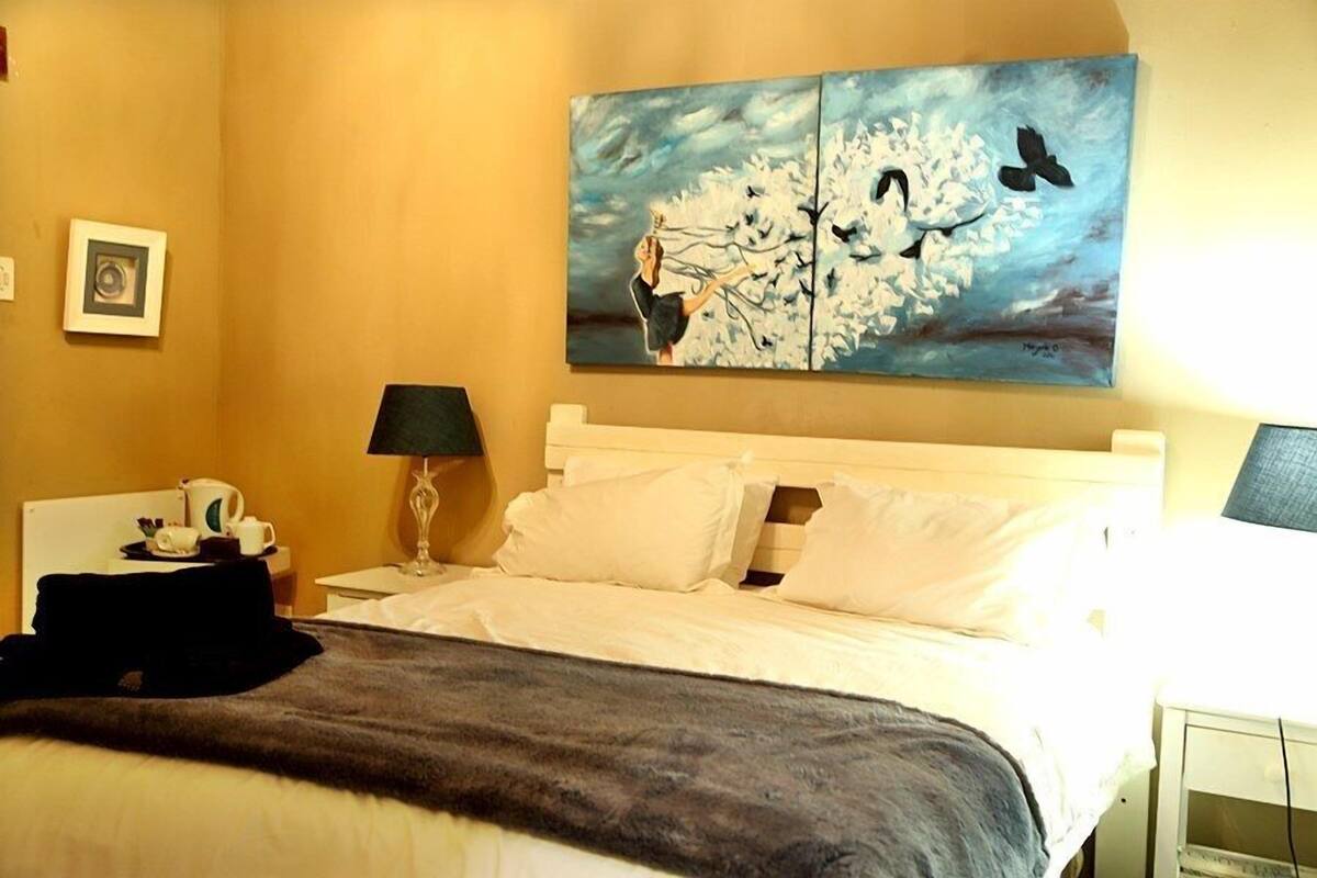A hotel room with a bed, a nightstand with a lamp, a painting on the wall, and a framed picture.