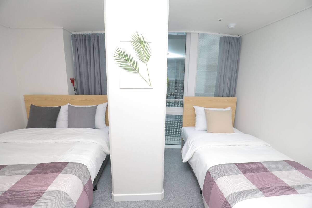 A hotel room with two beds, a large window with curtains, and a wall-mounted plant.