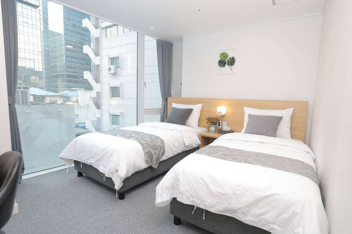 A hotel room with two beds, a large window with a city view, a small table with a lamp, and a wall-mounted plant.
