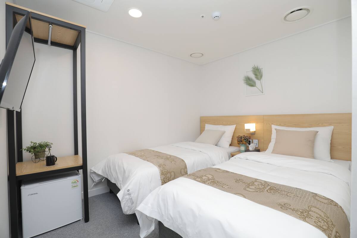 A hotel room with two beds, a desk with a chair, a small refrigerator, and a wall-mounted TV.