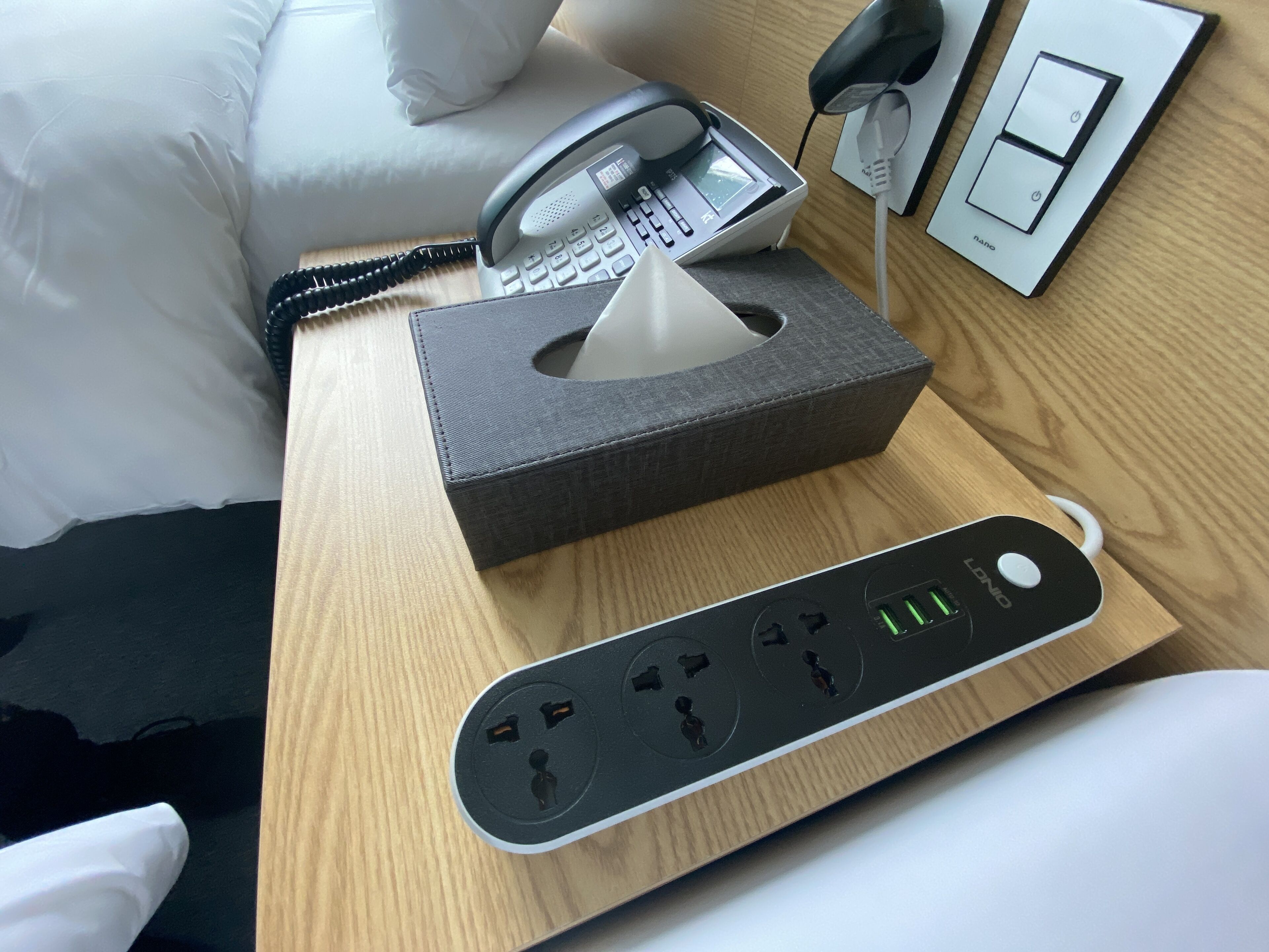 Et hotelværelse med et natbord, der har en telefon, en tissueske og en strømfordeler med flere stik og USB-porte.