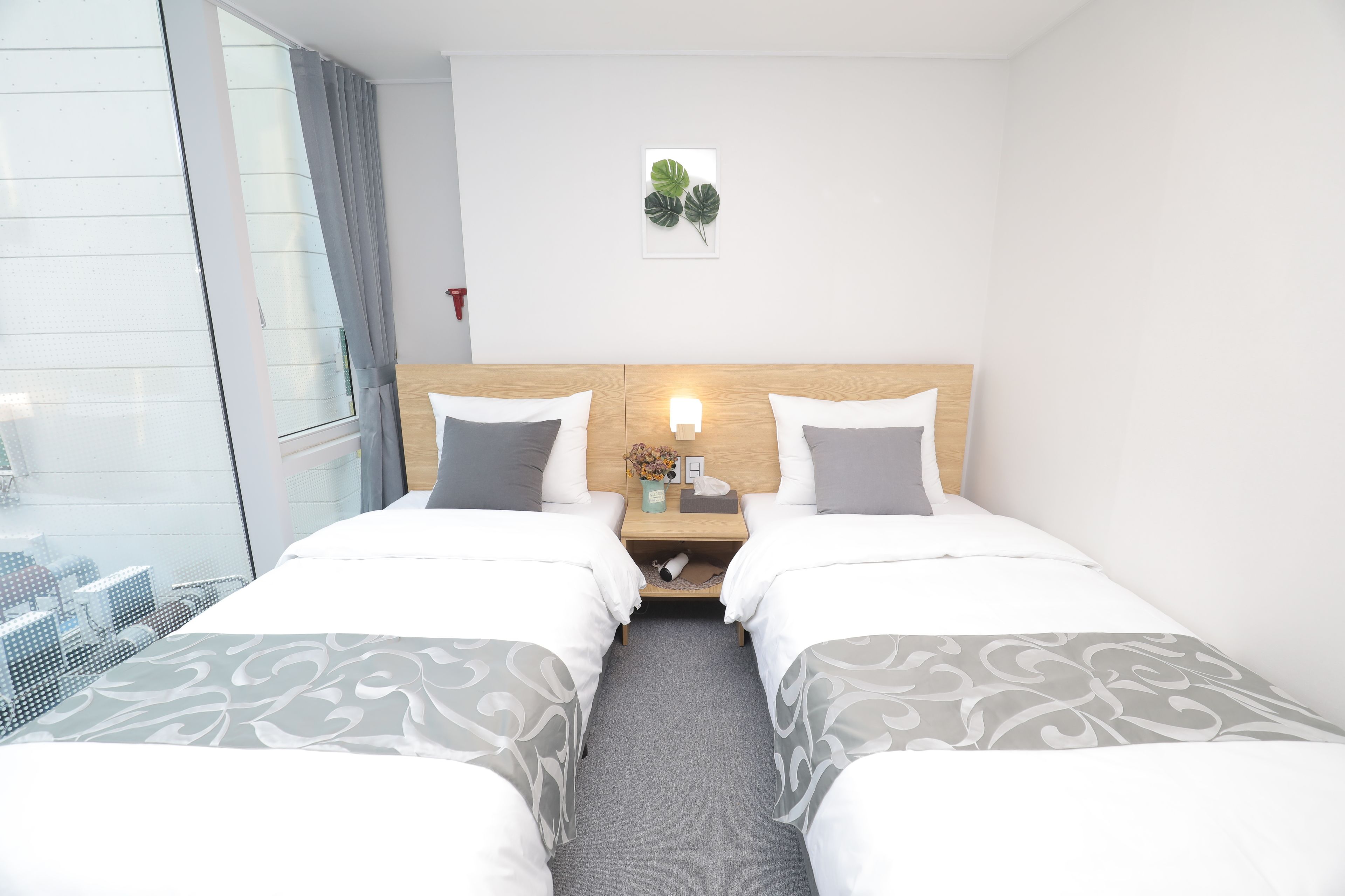 A hotel room with two beds, a bedside table with a lamp, a small plant, and a view of the city through a large window.