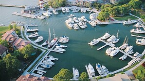 Vista aérea de un puerto deportivo con numerosos barcos amarrados en una distribución circular, rodeado de edificios residenciales y vegetación.