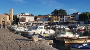 Un port de plaisance où sont amarrés différents types de bateaux, une église dotée d’un clocher, et des bâtiments abritant des boutiques et des restaurants.