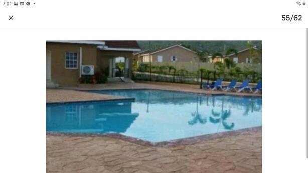 Une piscine avec une terrasse carrelée, un bâtiment doté d’une porte et de fenêtres, et une clôture avec des chaises.