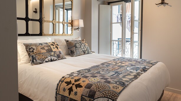 Ein ordentlich bezogenes Bett mit einem gemusterten Quilz und Kissen, ein an der Wand befestigter Spiegel und ein Fenster mit Blick ins Freie.