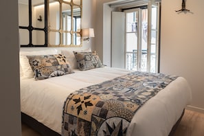 A neatly made bed with a patterned quilt and pillows, a wall-mounted mirror, and a window with a view of the outside. - Sitio dos Cavaleiros Apartments (Lisbon)