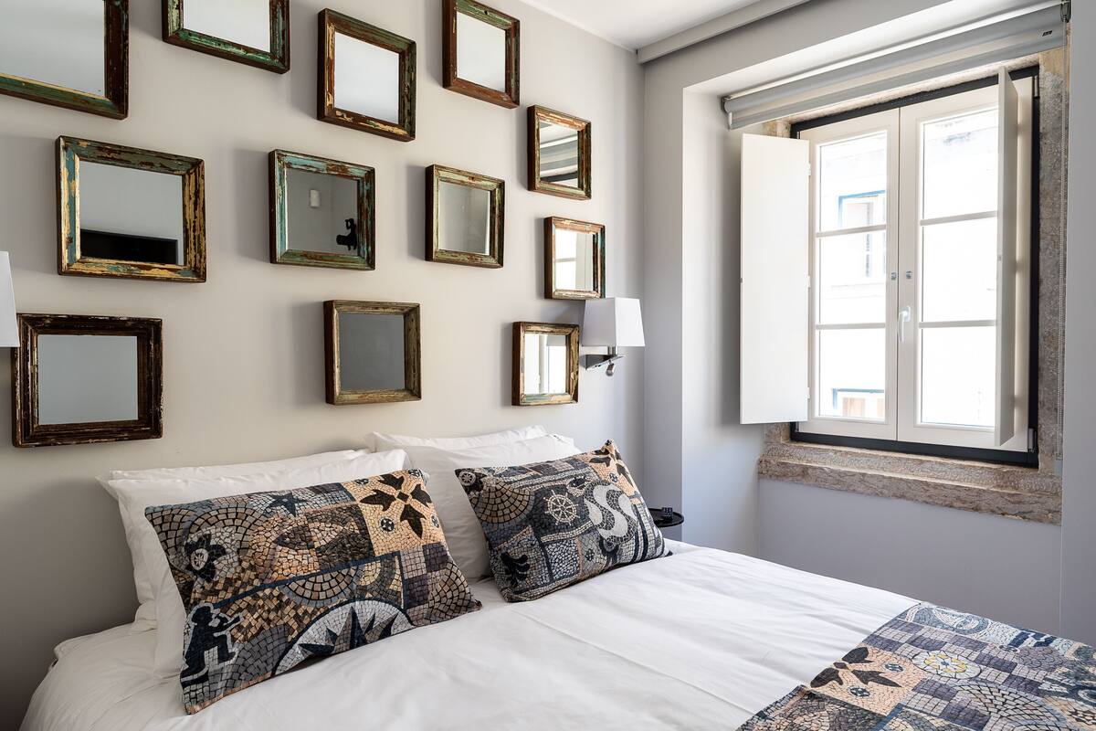 Un dormitorio con cama, varios espejos enmarcados en la pared y una ventana con vistas a edificios.