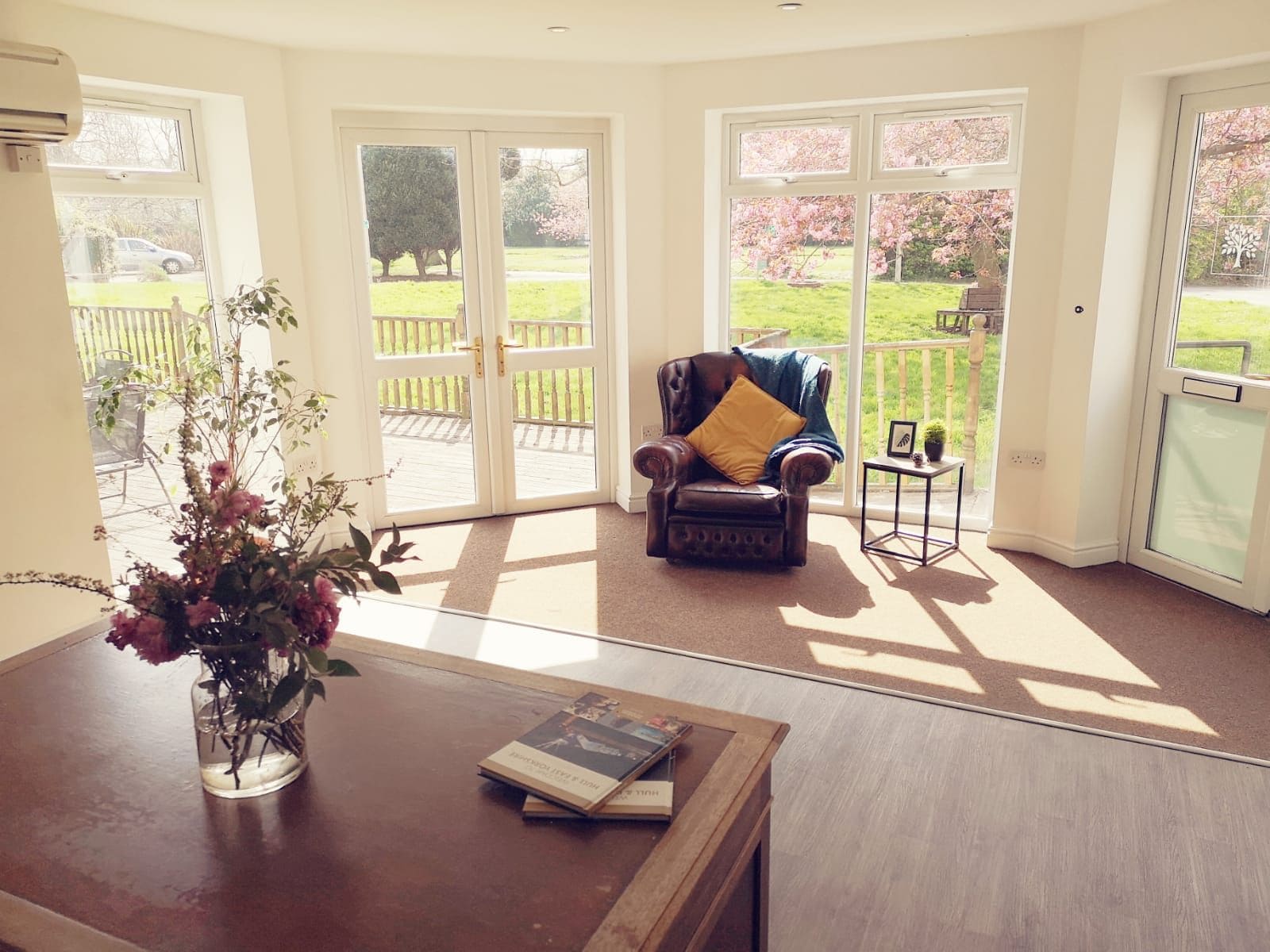 A room with a large window, a leather armchair, a small table with a vase of flowers, and a book on the table.