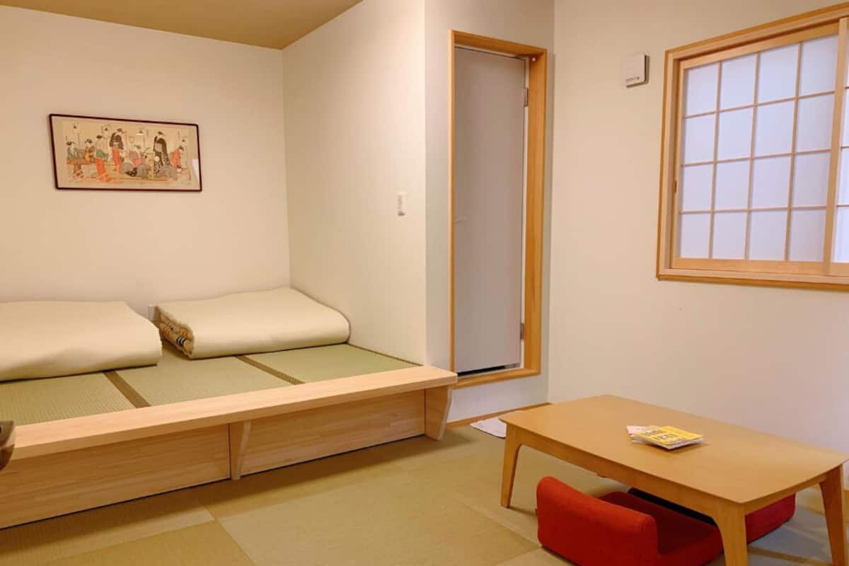 A room with a futon bed, a small table, a red bench, a window with a grid pattern, and a framed picture on the wall.