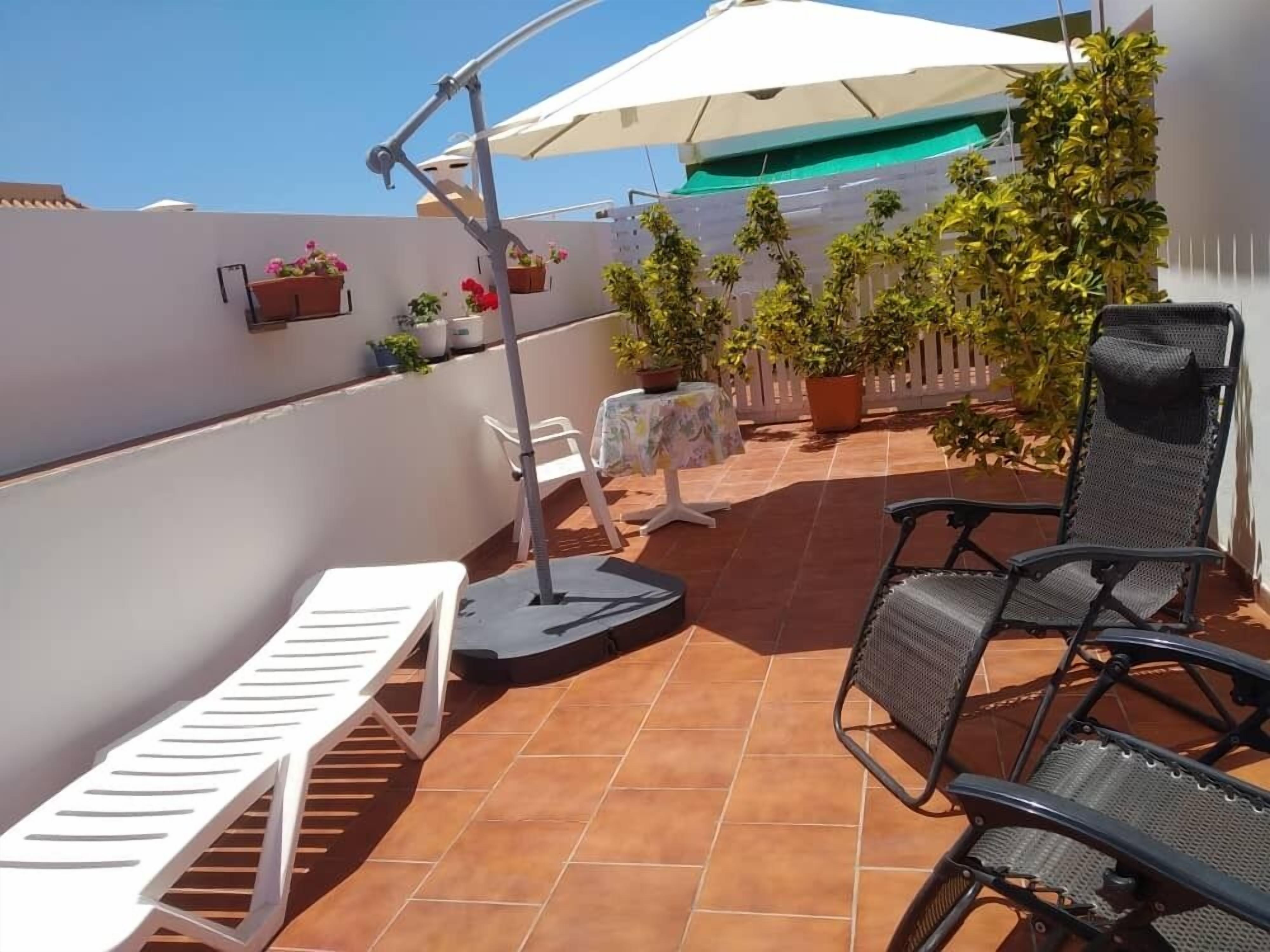A terrace with a white lounge chair, a black chair, and a table with a white tablecloth. There are potted plants and a white umbrella.