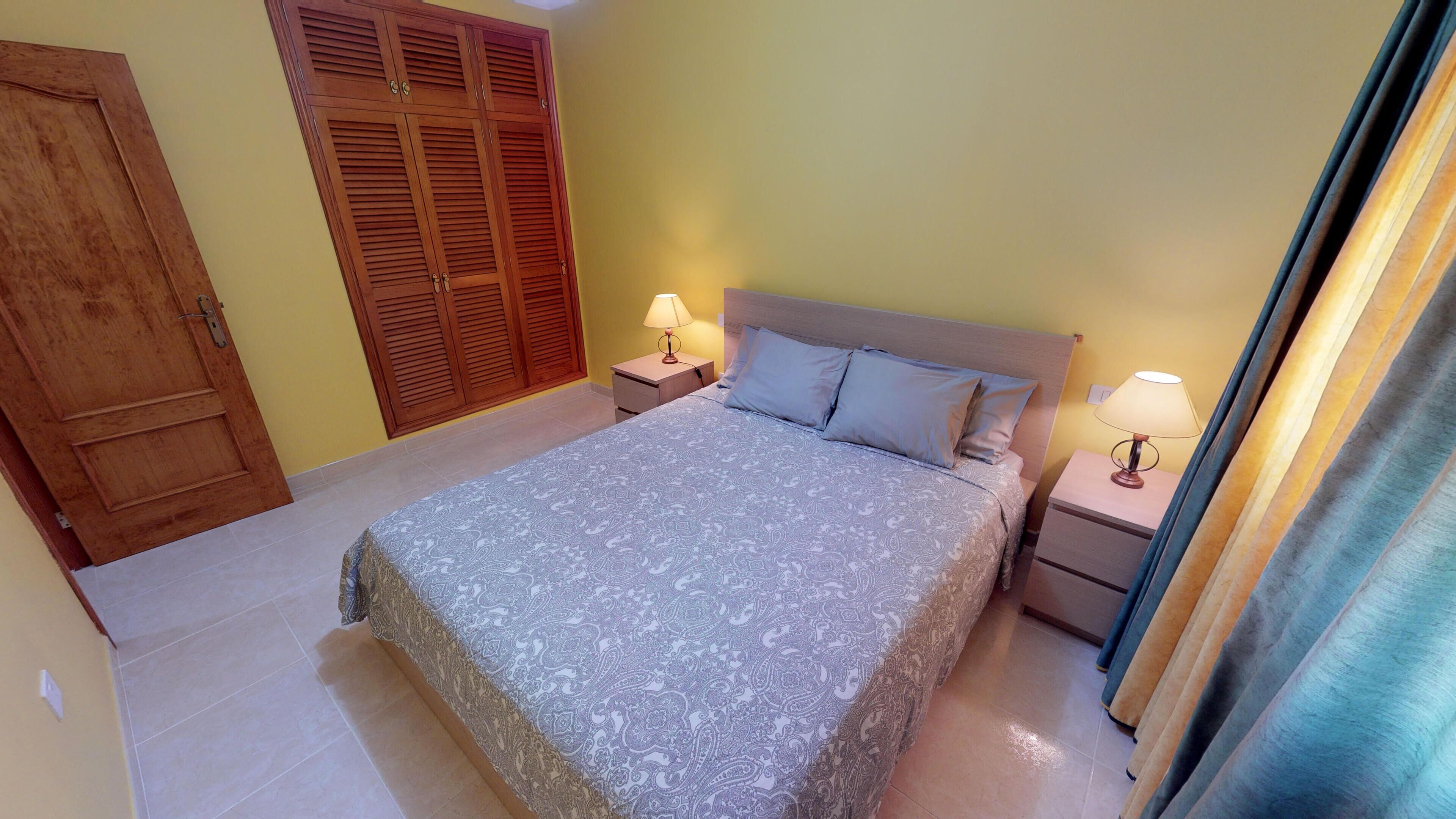 Un dormitorio con una cama, dos mesitas de noche con lámparas, un armario de madera y una cortina.