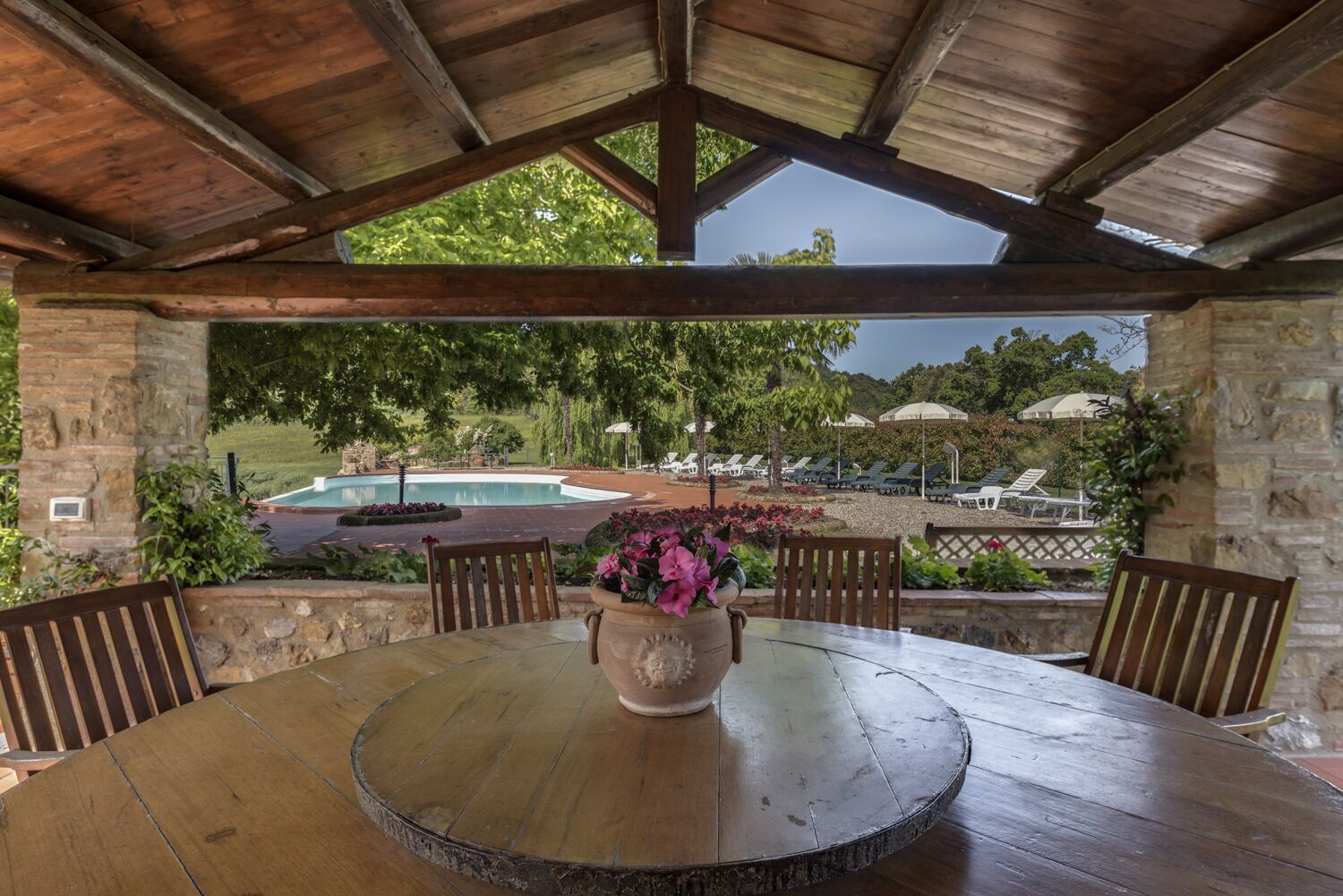 An outdoor patio with a round wooden table, stone walls, and a view of a swimming pool and lounge area.