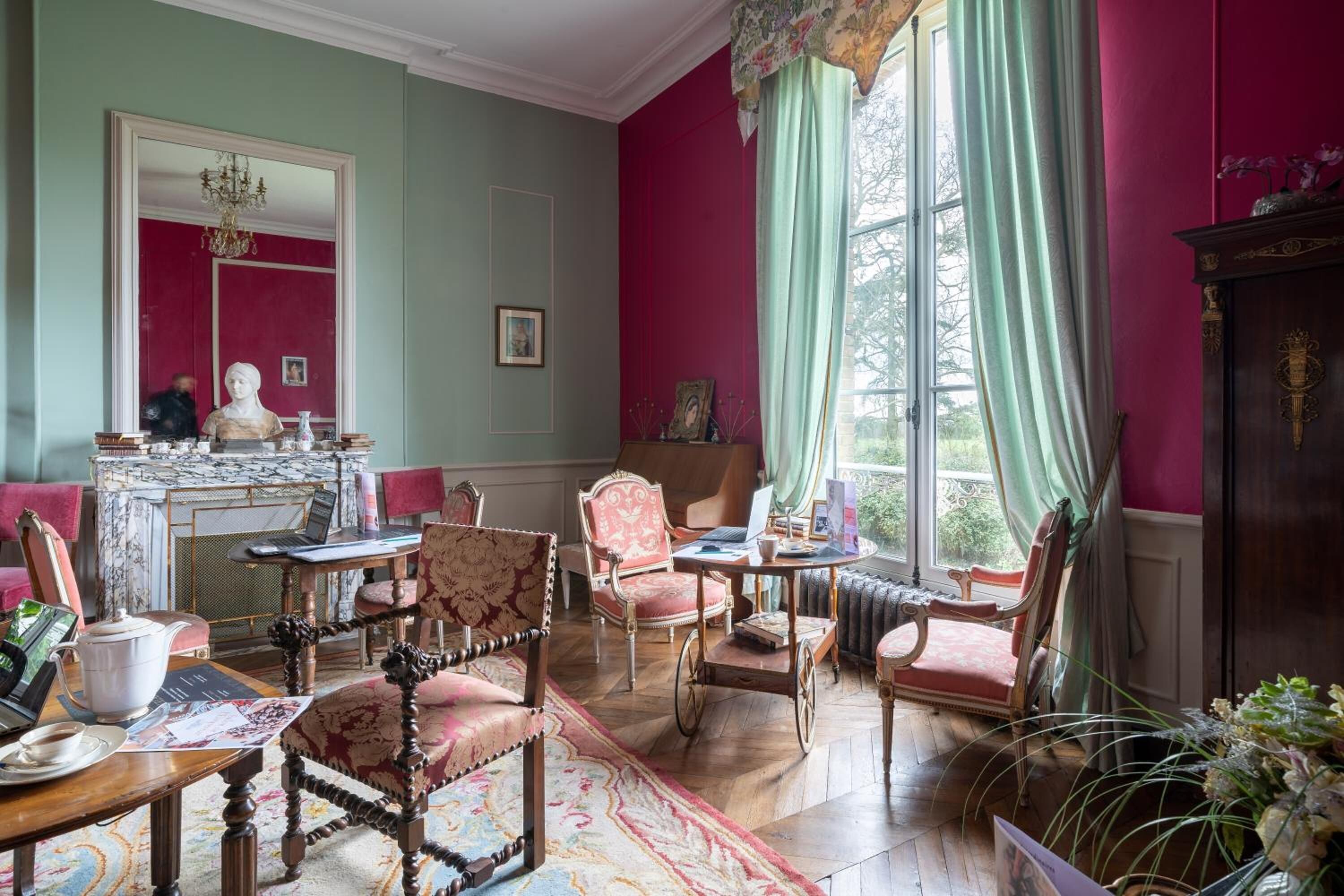 A room with a large window, a fireplace, and several antique chairs.