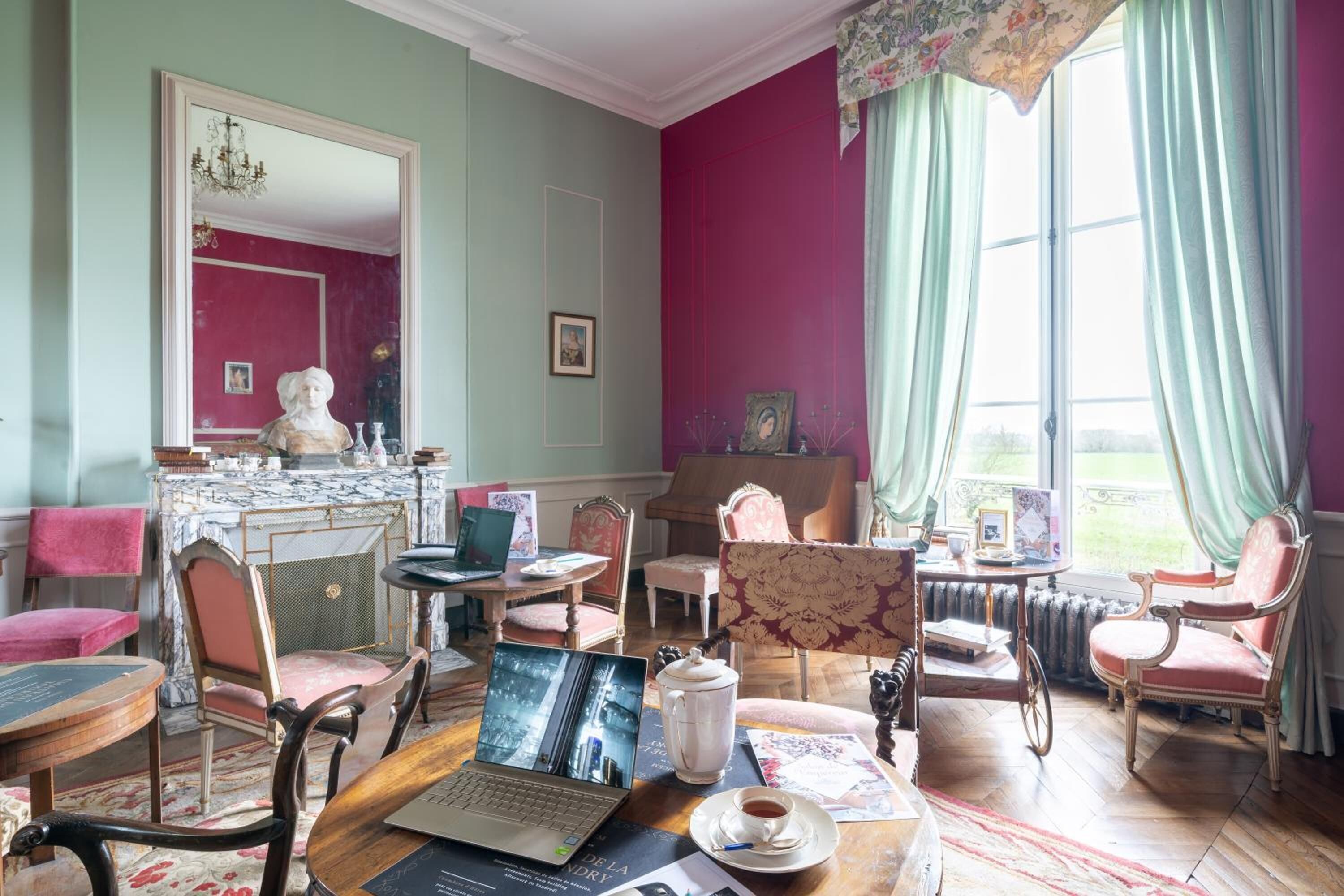 A room with a laptop, a teapot, and a cup on a wooden table. There are chairs and a sofa with a pink cushion. A large mirror and a window with curtains are visible.