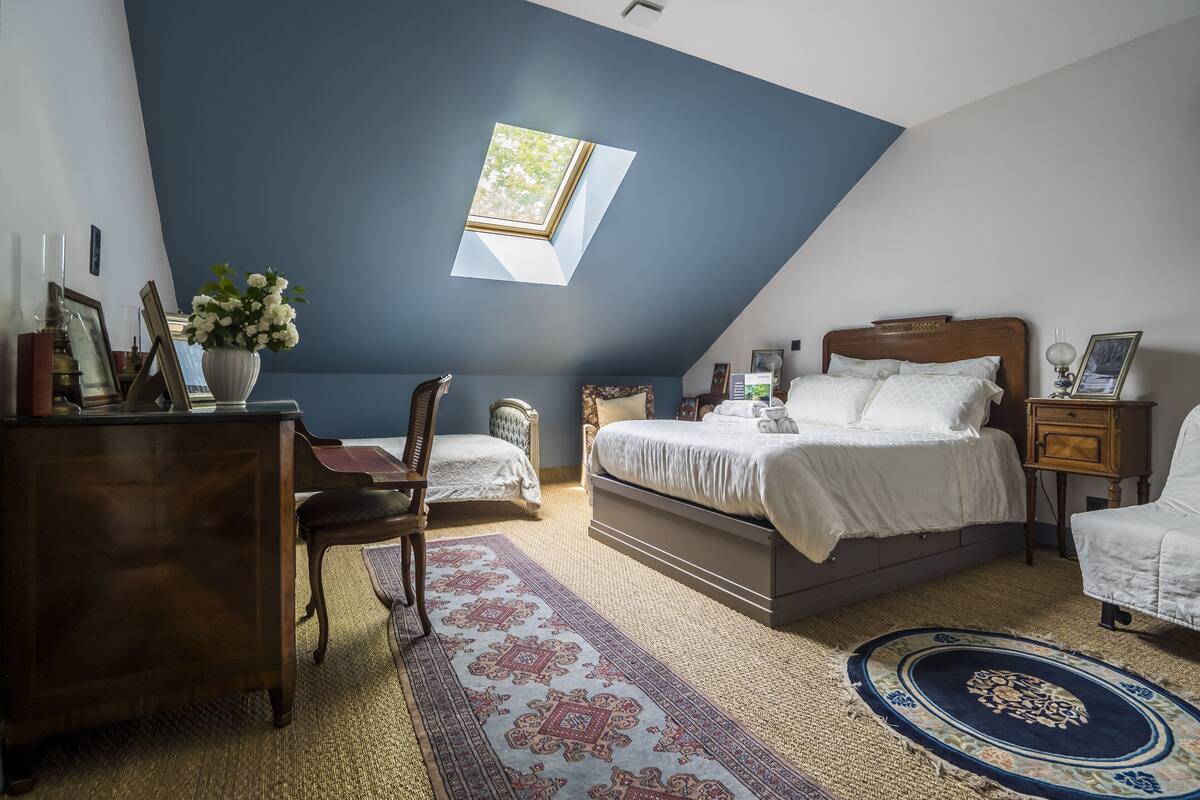 Ein Schlafzimmer mit schrägen Wänden, einem Oberlicht, einem Bett, einem Schminktisch, einem Sessel und einem Nachttisch.