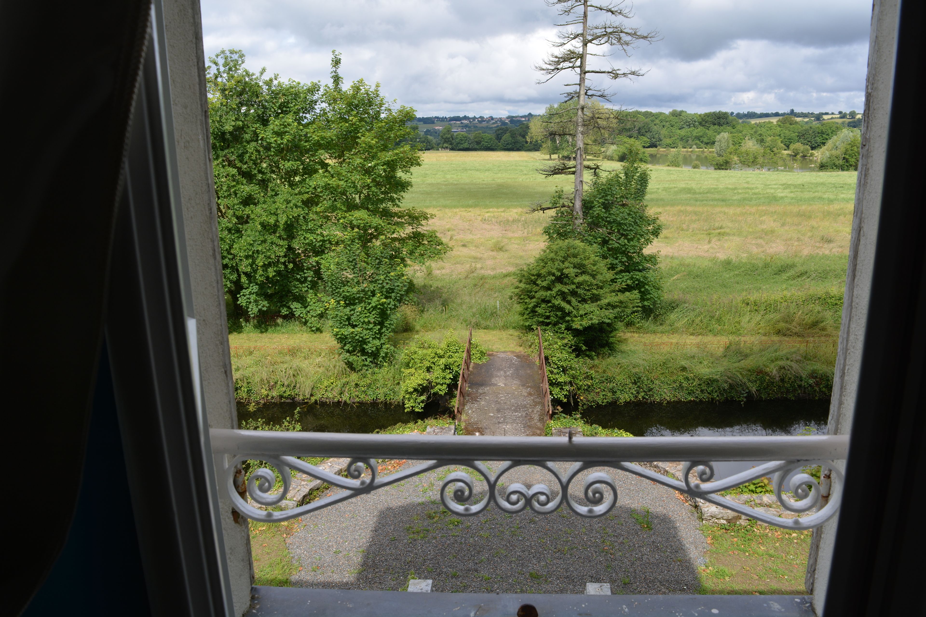 A view from a window with a metal railing, overlooking a grassy field, a narrow path, and a body of water.
