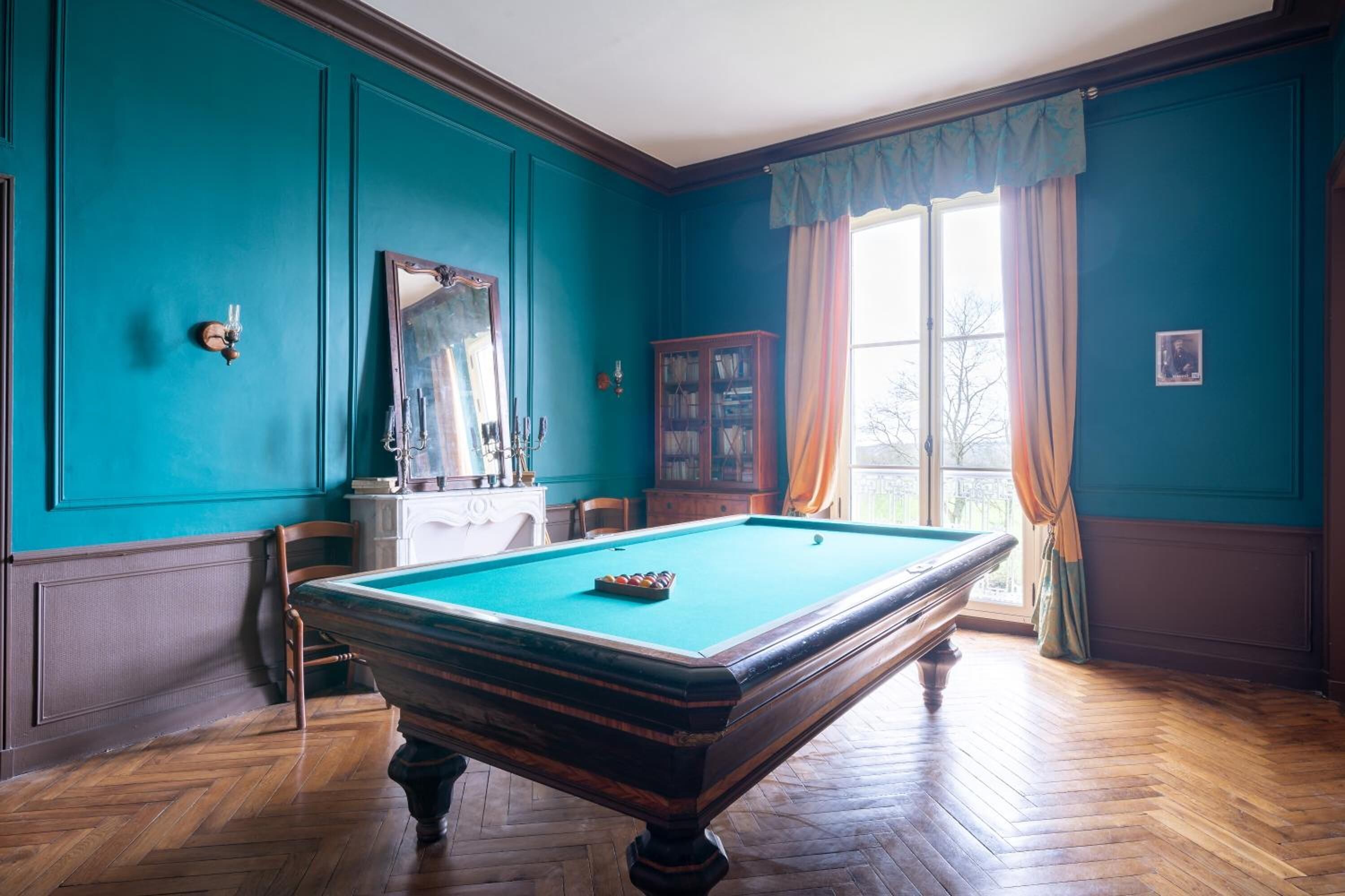 A billiards room with a pool table, wooden flooring, teal walls, a large mirror, and a window with curtains.