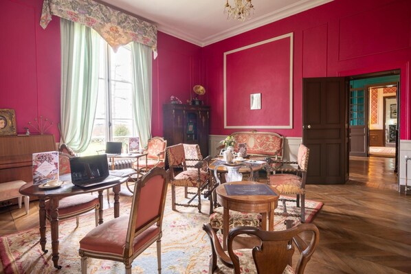 A room with a red wall, a wooden floor, and a large window with curtains. There are several chairs and a table with a vase of flowers.