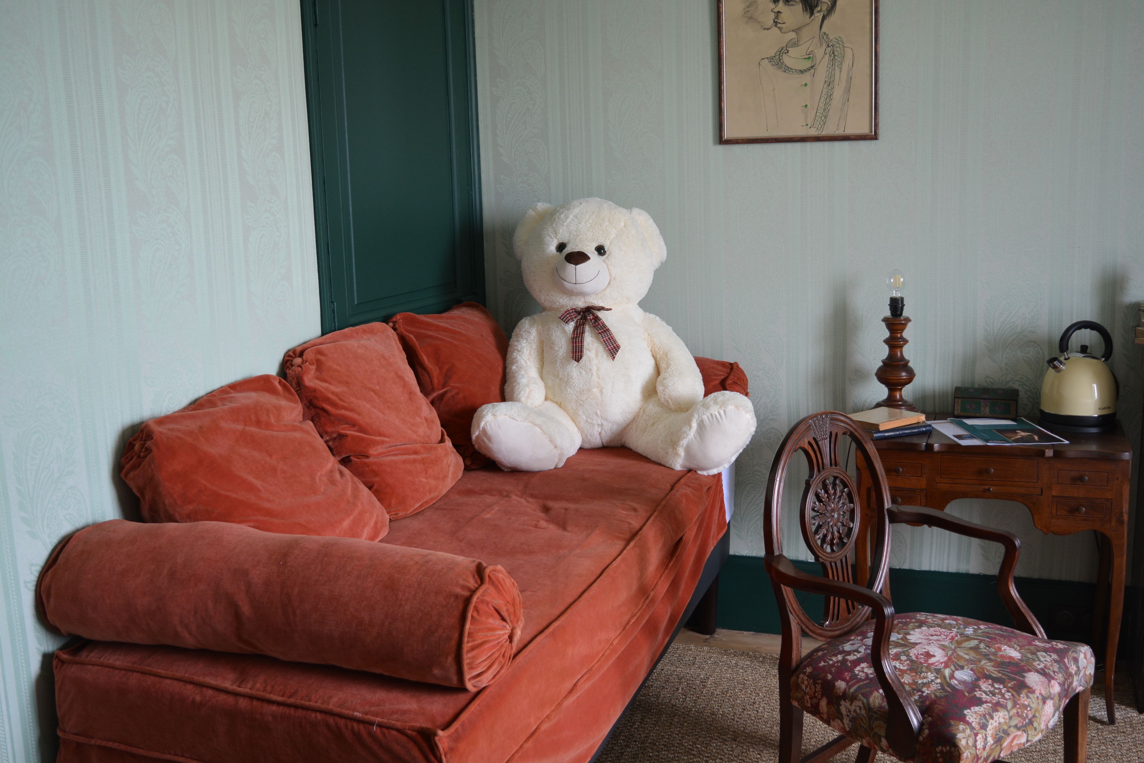 Ein Zimmer mit einem roten Sofa, einem großen weißen Teddybären, einem Holzsessel und einem Schreibtisch mit einer Lampe.