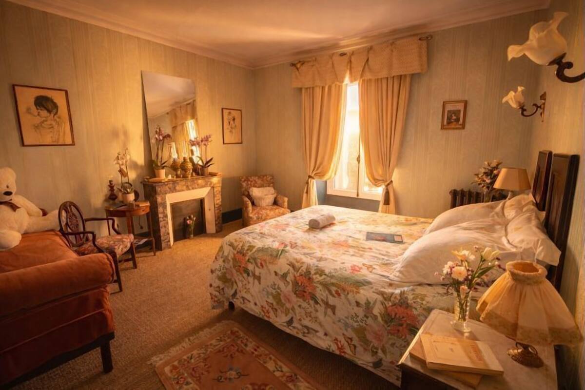 Ein Schlafzimmer mit einem Bett, einem Sessel, einer Kommode, einem Spiegel, einem Fenster mit Vorhängen und einem Kamin.