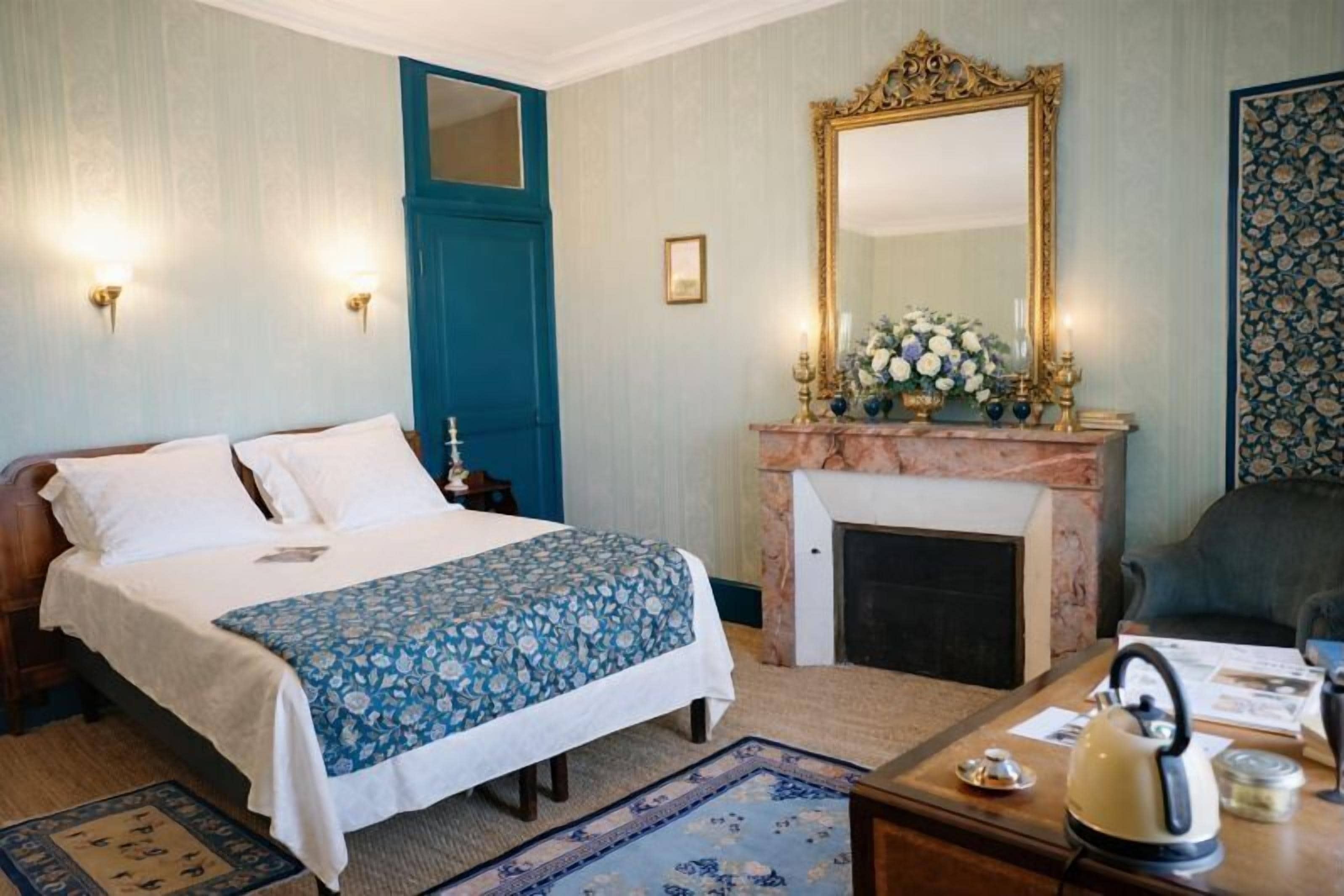 A bedroom with a bed, a fireplace, a mirror, and a desk with a kettle.