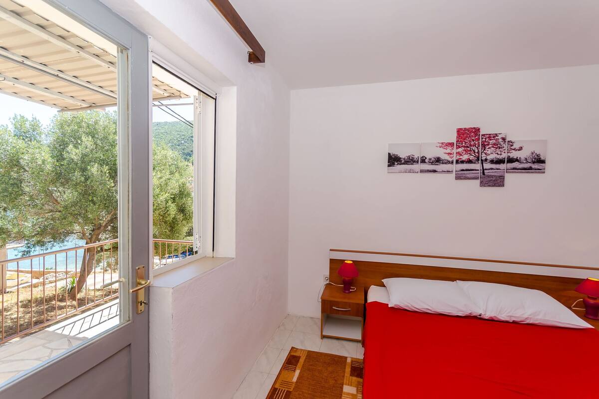 A bedroom with a bed, a nightstand, a lamp, a window with a view of a beach, and a wall with three framed pictures.