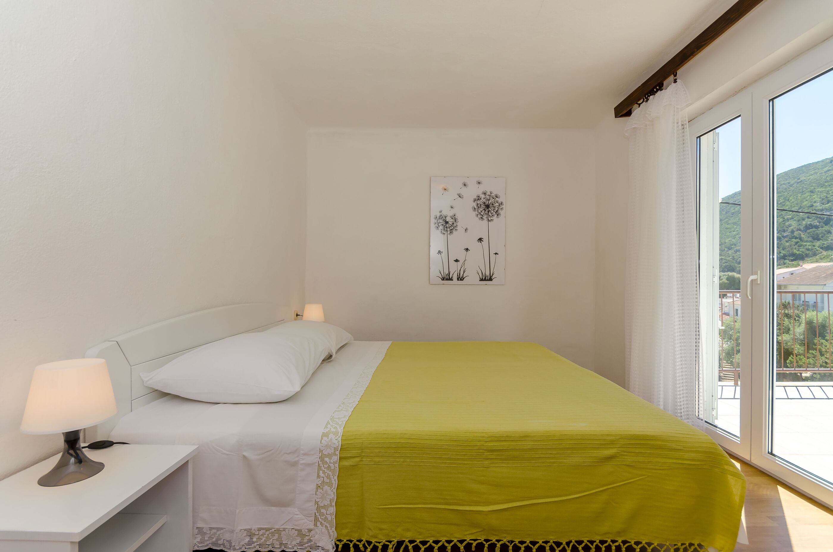 A bedroom with a bed, nightstand, lamp, and a view of greenery through a window.
