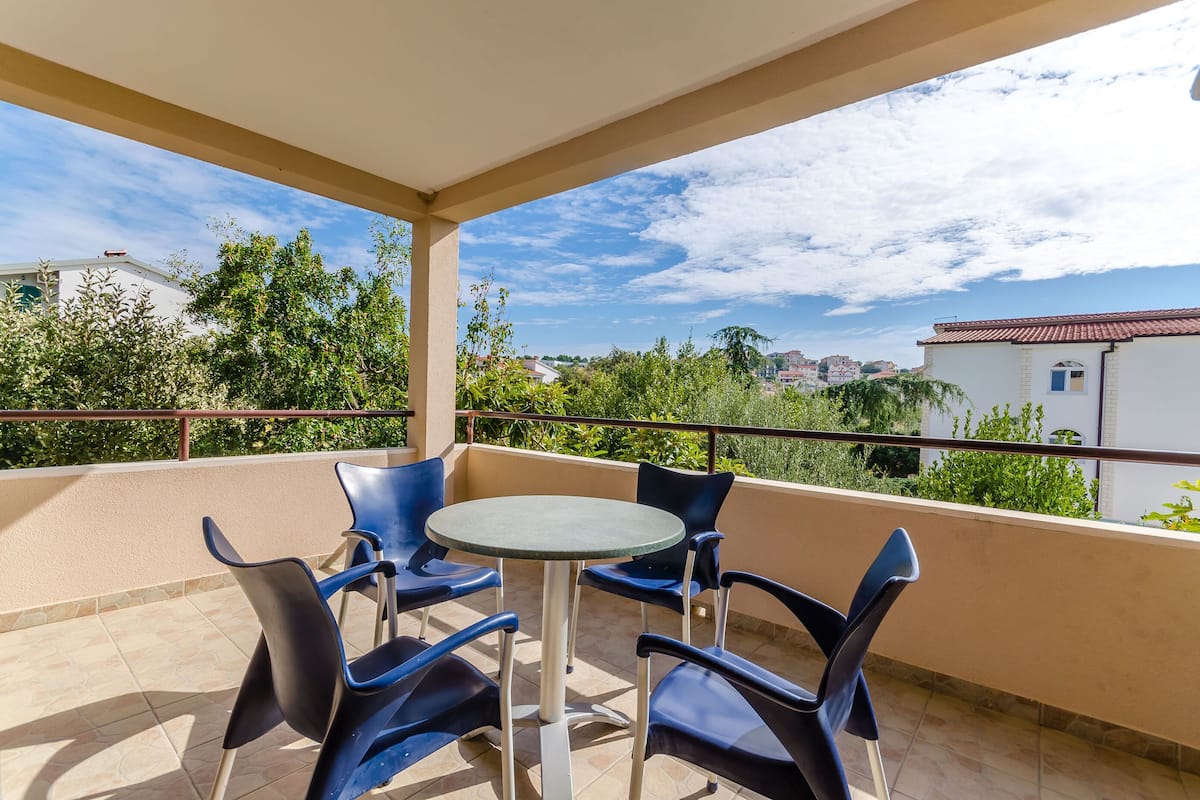 Un balcon avec une table ronde et quatre chaises, donnant sur un quartier résidentiel bordé d’arbres et de bâtiments.