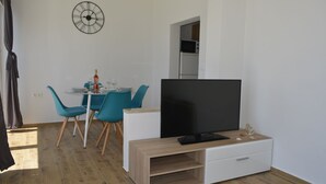 En moderne stue med et fladskærms TV på et træstel, et spisebord med blå stole, og et køkken der kan ses gennem en åben dør.