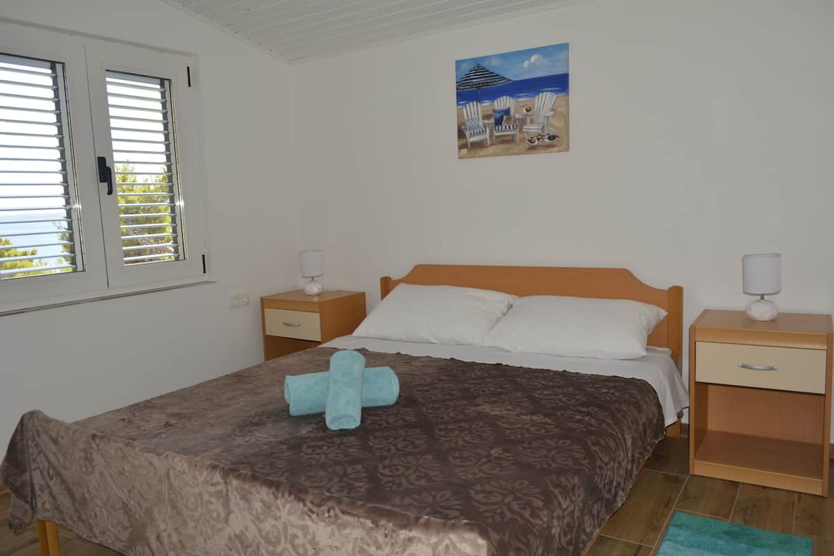 A bedroom with a bed, a nightstand, a bedside table, a window with blinds, and a beach-themed painting on the wall.