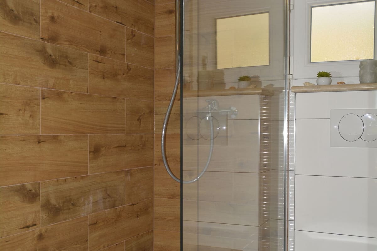 A modern bathroom with a glass-enclosed shower, wooden wall tiles, a white toilet, and a small sink with a mirror above it.