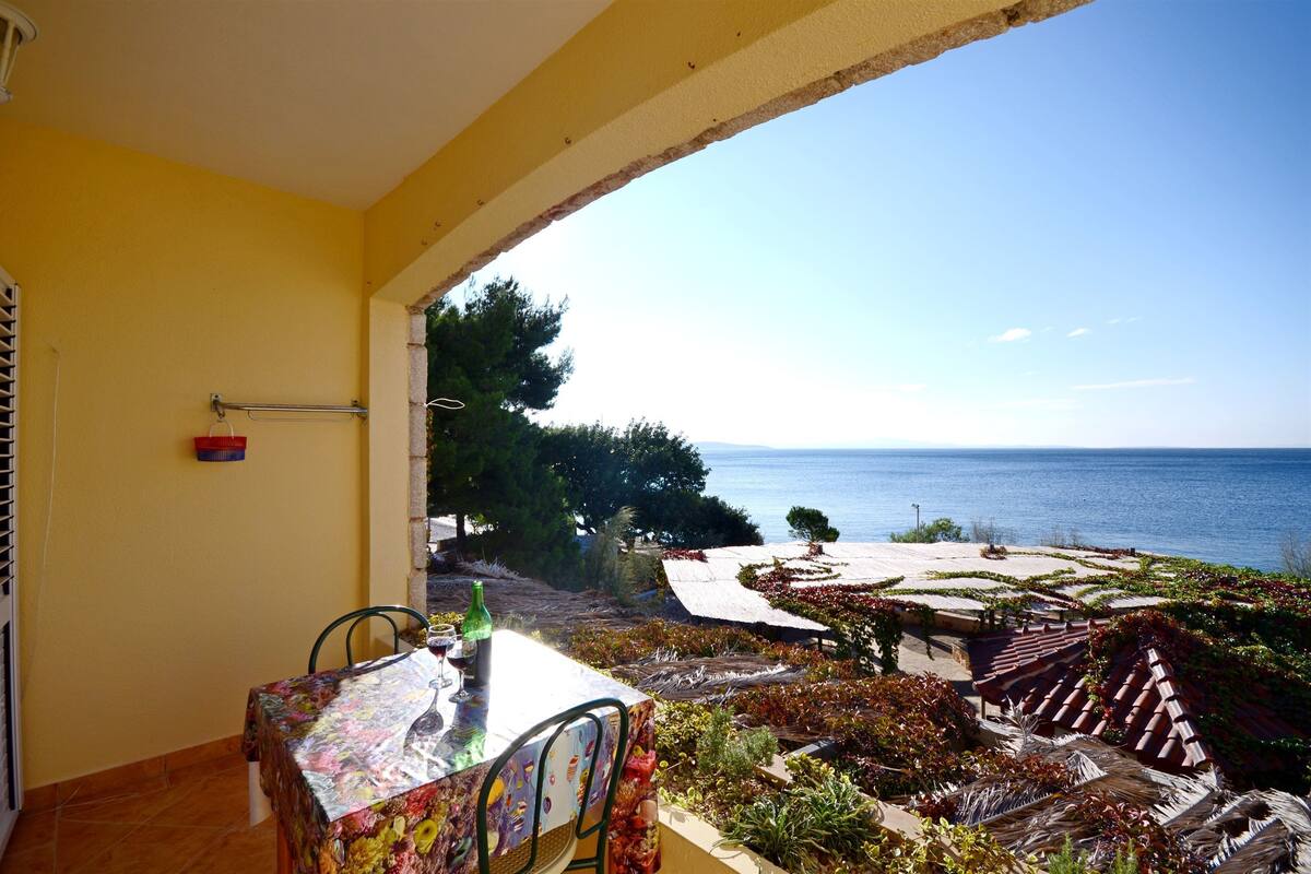 A balcony with a table and chairs, overlooking a coastal landscape with terraced gardens and the sea.