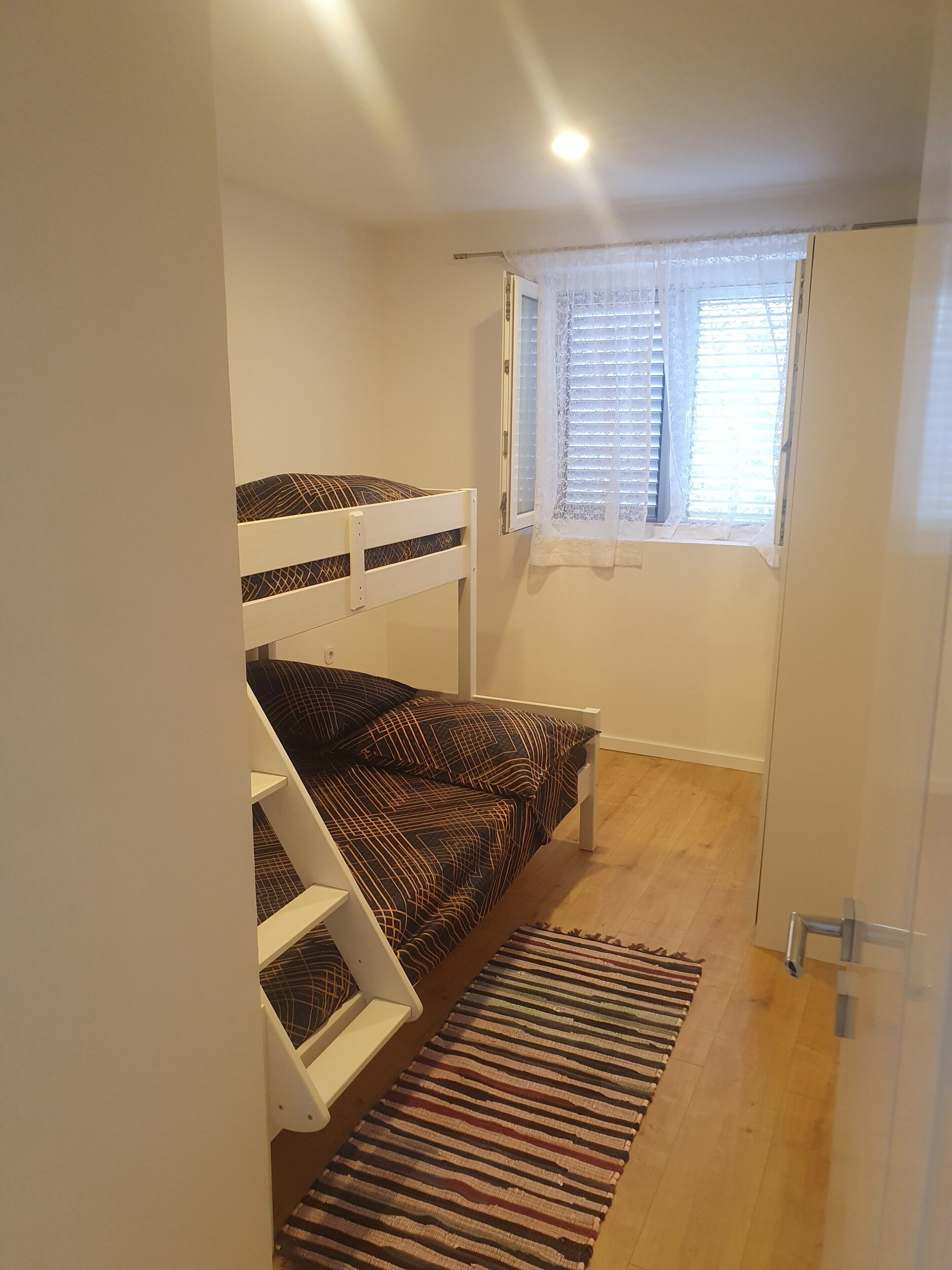 A small, compact room with a bunk bed, a window with blinds, a wardrobe, and a striped rug on the floor.