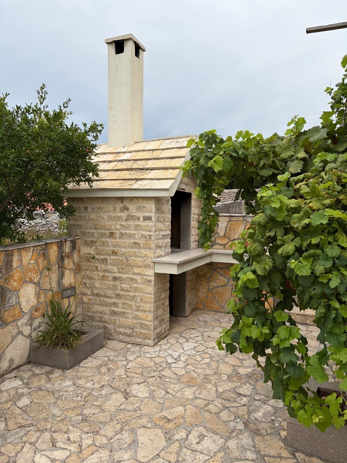A stone-built outdoor kitchen with a chimney, surrounded by stone walls and potted plants.