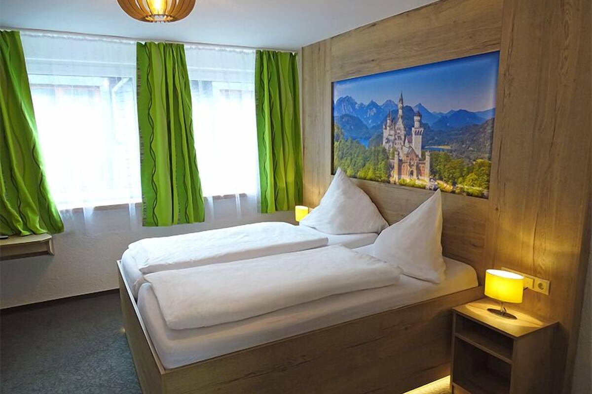Un lit double avec du linge de lit blanc, des rideaux verts, une tête de lit en bois et un tableau représentant un château.