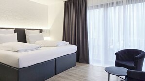 Une chambre d’hôtel moderne avec un grand lit, un fauteuil sombre et une vue sur l’extérieur à travers des rideaux légers.