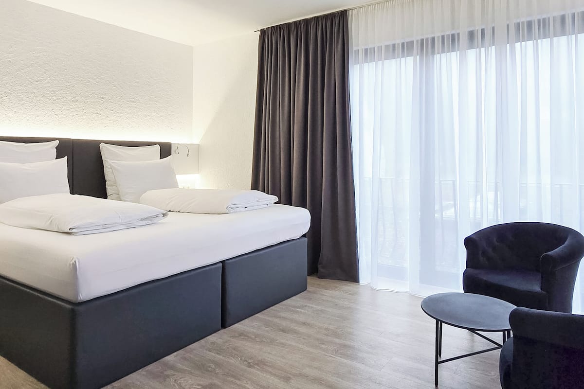 Une chambre d’hôtel moderne avec un grand lit, un fauteuil sombre et une vue sur l’extérieur à travers des rideaux légers.