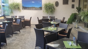 An outdoor seating area with wicker furniture and glass tables, surrounded by potted plants and a cityscape painting.