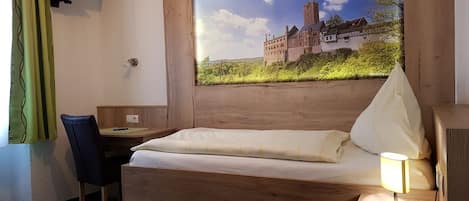 A hotel room with a wooden bed, a desk, a chair, a TV mounted on the wall, and a large window with a castle view.