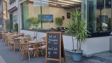 An outdoor cafe with wicker tables and chairs, a chalkboard menu, and potted plants.