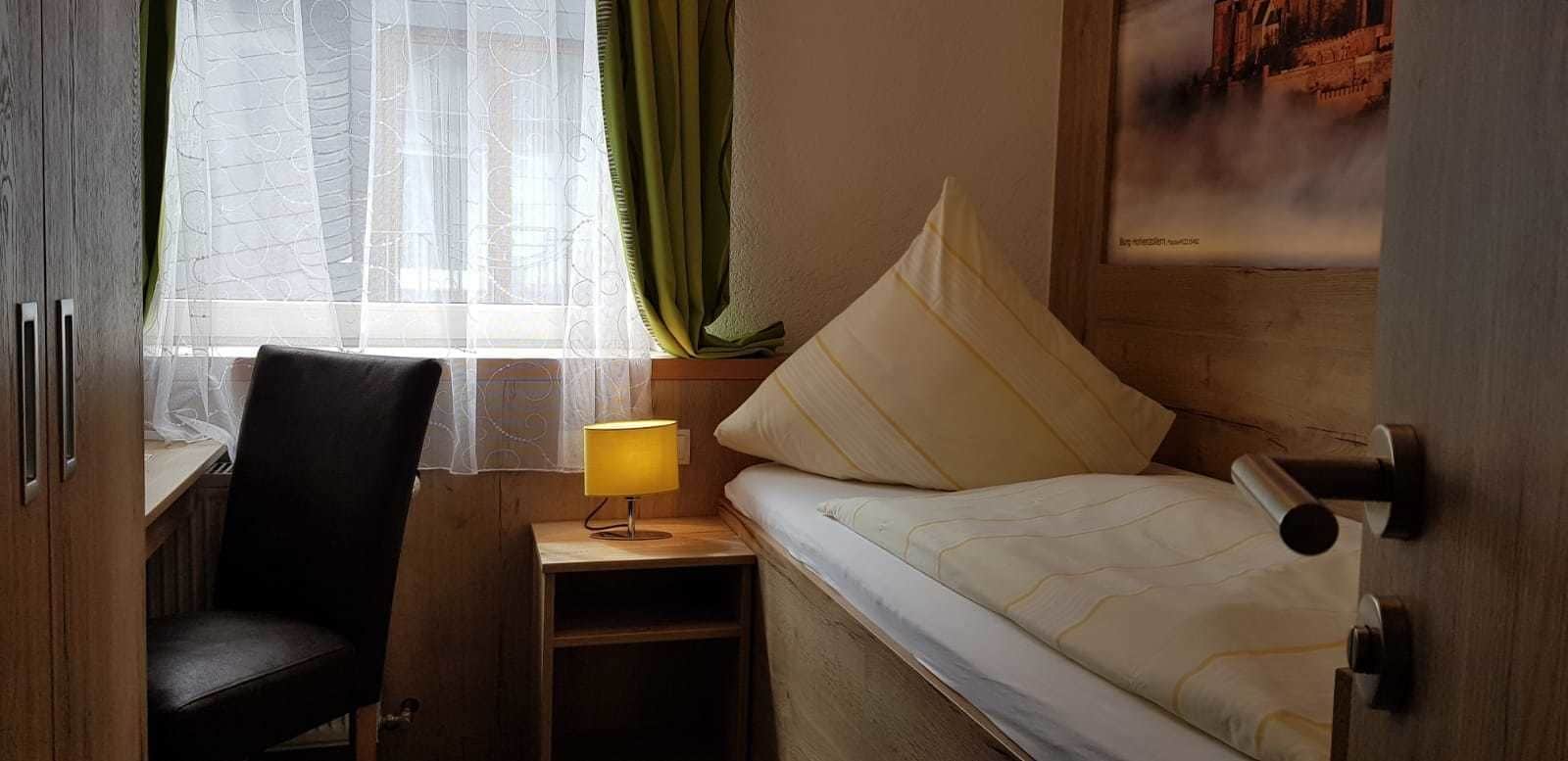 Ein kleines Hotelzimmer mit Bett, Stuhl, Nachttisch mit Lampe und Fenster mit transparenten Vorhängen.