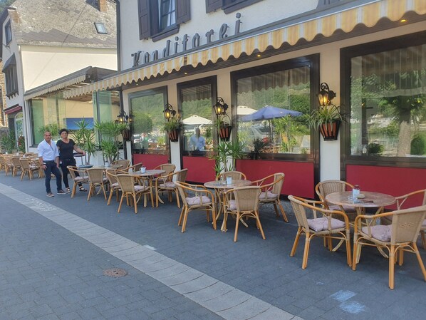 Un café al aire libre con mesas y sillas de mimbre, ventanas amplias y plantas en macetas.