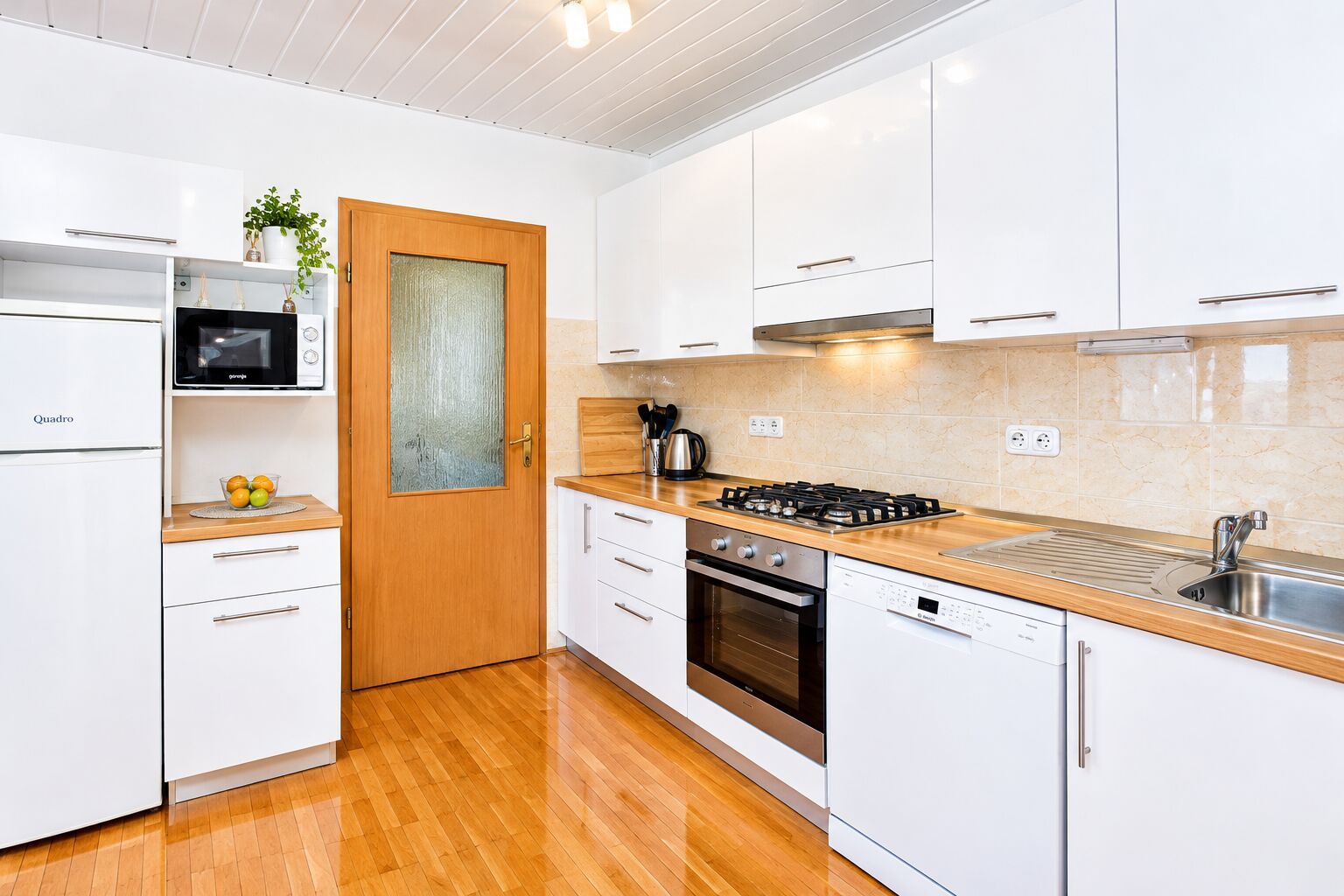 A modern kitchen with white cabinets, a wooden door, a stove, and a dishwasher.