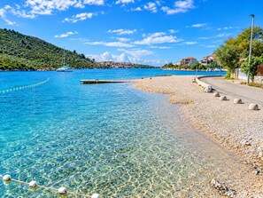 A clear coastal view with a pebble beach, a boat dock, and buildings in the background. - Apartment Bosiljka (Rogoznica)