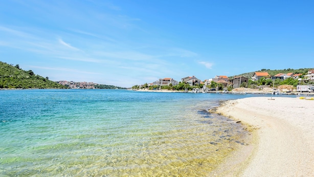 Una playa costera con agua cristalina, orilla de arena y edificios residenciales al fondo.